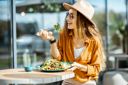 Stylish young woman eating healthy salad on a restaurant terrace, feeling happy on a summer day
