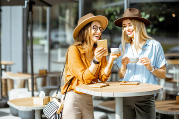 Two female best friends spending time together on the cafe terrace, feeling happy standing with coffee and phone during a summer day