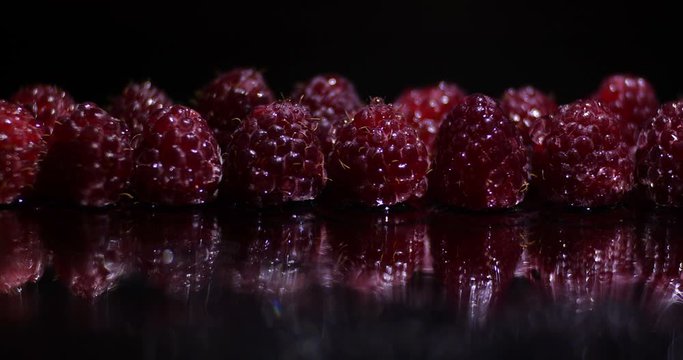 
Close up macro studio  dolly shot movement  along a row of fresh raspberries in water droplets with bokeh on a black background with reflection, dramatic light changing . 4K
