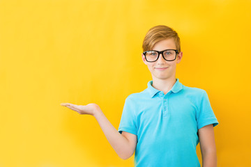 Positive handsome teen boy stands with open palm. Copy space and yellow background