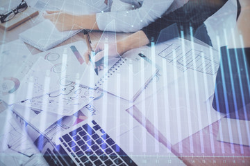 A woman hands writing information about stock market in notepad. Forex chart holograms in front. Concept of research. Double exposure