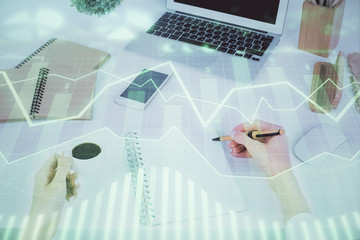 Hands writing information about stock market in notepad. Forex chart holograms in front. Concept of...