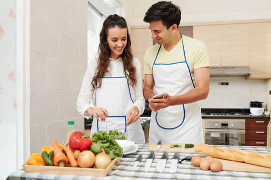 Cheerful young man showing funny pictures on smartphone screen to girlfriend who is mixing healthy salad for dinner