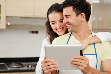 Pretty smiling young woman hugging her boyfriend from behind and pointing at screen of digital tablet in his hands when that are standing in kitchen