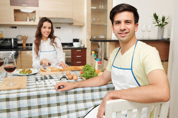 Portrait of handsome young man in apron sitting at dinner table and looking at camera, his wife cutting bread in background