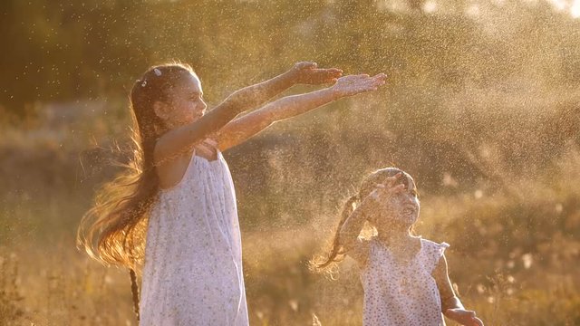 Two little pretty sisters, four and seven years old having fun joyfully jumping in flying spray of water  in warm bright sunset rays  on a sunny summer evening. Slow motion  200 fps 