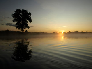 Fototapeta na wymiar foggy blured orange sunrise at a river, the burning sky is reflected in calm water , dark tree silhouettes, Salaca river, Burtnieks lake, Latvia