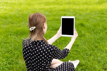 Young blonde girl in a summer dress makes selfie on a tablet while sitting on green grass in a park.