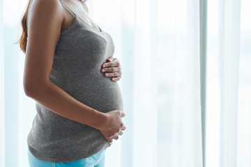 Cropped image of young woman in grey top touching her baby bump when standing against big window