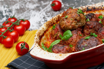 meatballs with tomato sauce and decorated with basil leaf, served in white and red pan on grey background. next to the pan near fresh tomatoes, spaghetti. Closeup.