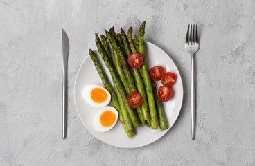 Baked green asparagus, fresh tomatoes and boiled eggs with herbs on a plate on a concrete background