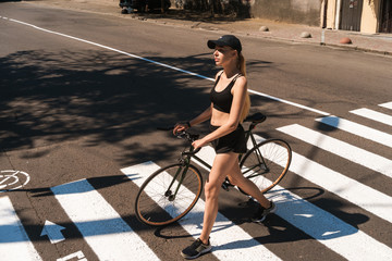 Image of athletic blonde woman dressed in sportswear and cap walking with bicycle at pedestrian...
