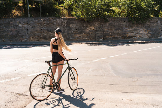 Image of pretty blonde woman dressed in sportswear and cap walking with bicycle at city road