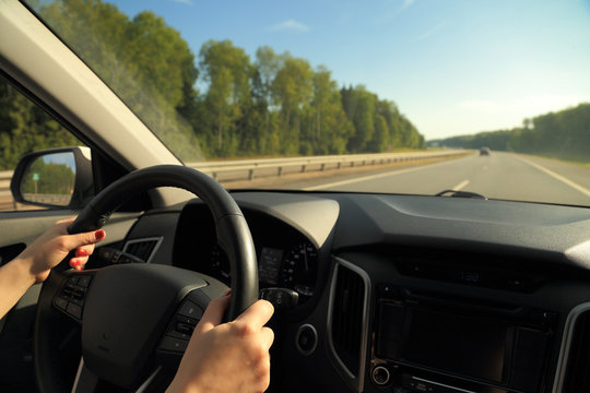 Female hands, lit by sunlight, hold the steering wheel of a car while driving on the road.