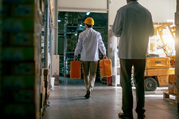 Obraz premium Rear view of strong Caucasian storage worker dressed in white uniform and with yellow helmet carrying canisters with oil while supervisor watching him. Storage interior.