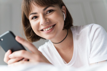 Smiling pleased young beautiful woman lies indoors at home on bed listening music with bluetooth...