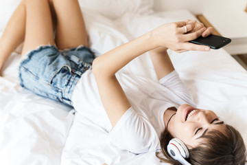 Obraz na płótnie Canvas Pleased cheery young beautiful woman indoors at home on bed listening music with headphones using mobile phone.