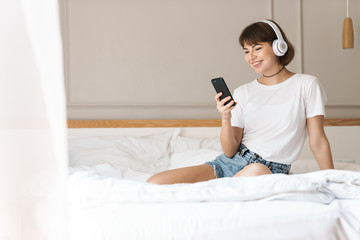 Pleased cheery young beautiful woman sitting indoors at home on bed listening music with headphones using mobile phone.