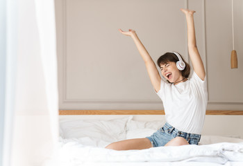 Cheery young beautiful woman sitting indoors at home on bed listening music with headphones.