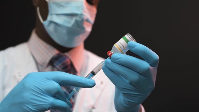 African American doctor drawing vaccine from vial into a syringe on a black background wearing white coat and blue gloves with mask