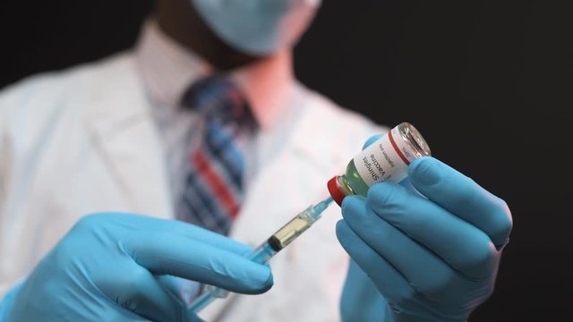 African American doctor drawing vaccine from vial into a syringe on a black background wearing white coat and blue gloves with mask