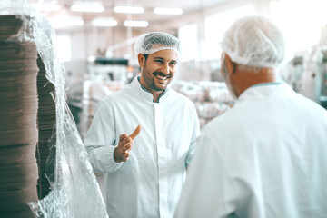 Two cheerful Caucasian food plant workers in white uniforms and with hairnets standing and talking...