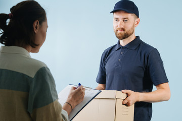 Woman is accepting a box delivery from a courier, signing paper form with pen
