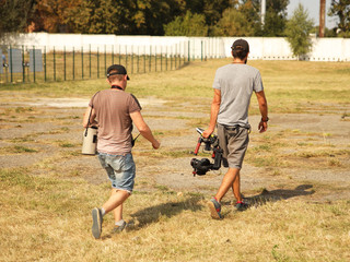 Bloggers videographer and photographer go with the camera on the stabilizer on a fenced field with grass. Modern technologies for the production of video content. Travel vlogers with gimbal in search