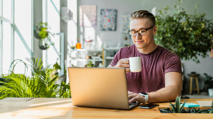 Front view shot of smiling young businessman drinking coffee while using laptop in office