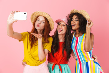 Happy amazing young three multiethnic girls friends posing isolated over pink wall background take a selfie by mobile phone.