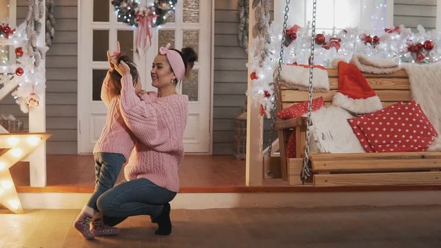 Beautiful mother and daughter in pink pullovers at Christmas day. Mother tickling her little daughter in slow motion. Happy mother having fun with her kid daughter at Christmas porch. Full length shot