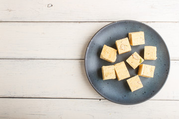 traditional indian candy soan papdi in a blue ceramic plate with almond and pistache on a white wooden background. top view