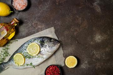 Raw fish dorado with lemon slices, herbs thyme, salt and pepper on dark background. Healthy food and diet concept. Top view, copy space. Ingredients for cooking fish
