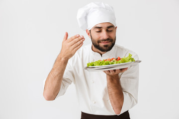Image of caucasian chief man in cook uniform smelling dish while holding plate with vegetable salad