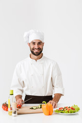 Image of handsome chief man in uniform smiling and cooking vegetable salad on wooden cutting board