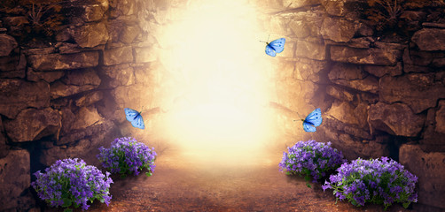 Photo background with magical trail leading through stone dungeon cave towards mystical glow, fantastic bluebells campanula flowers, flying blue butterflies. Fairytale tranquil fantasy scene