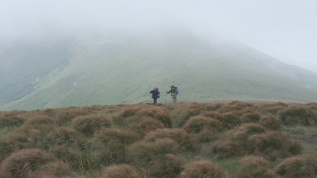 Two travelers climb the mountain on a hiking trail with trekking poles. Traveling with a backpack in bad weather with a strong wind, gale. Strong wind in the mountains.