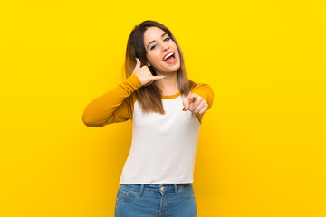 Pretty young woman over isolated yellow wall making phone gesture and pointing front