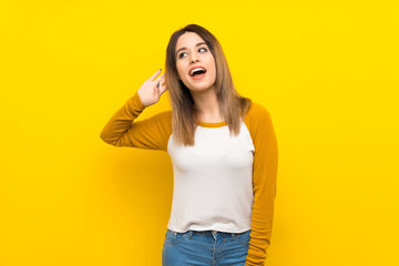 Pretty young woman over isolated yellow wall listening to something by putting hand on the ear