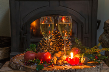 Cozy Christmas holiday still life. Two glasses of champagne, burning candle, apples, pine cones on the tree stump in front of the wooden stove
