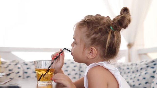 Cute little girl drinks juice using drinking straw at the cafe on seashore. Video shot of people on vacation in 4K definition.