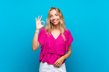 Fototapeta premium Blonde young woman over isolated blue background showing ok sign with fingers