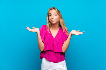 Blonde young woman over isolated blue background having doubts while raising hands