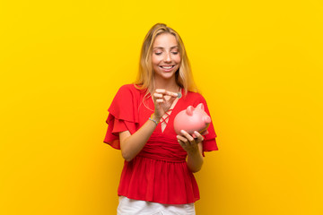 Blonde young woman over isolated yellow background holding a big piggybank