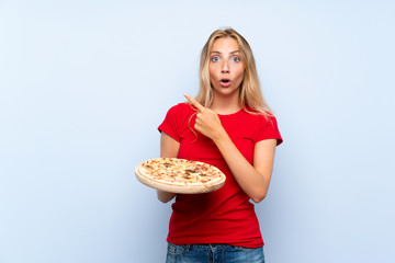 Young blonde woman holding a pizza over isolated blue wall surprised and pointing side