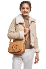 Medium full shot of a young chestnut European lady with a shoulder bag is wearing white casual trousers, a beige turtleneck and a short cream jacket with a fur trim. The bag is made of smooth sandy  © RedUmbrella&Donkey