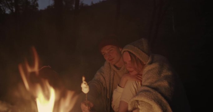 A young couple sits at night in the forest, embracing under a common plaid in front of a bonfire, talking and laughing.