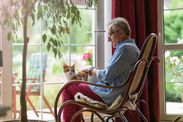 Senior man sitting in a rocking chair with his cat in his lap - obrazy, fototapety, plakaty