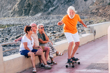 group of seniors and mature people at the beach have fun looking at old man riding a skateboard and...