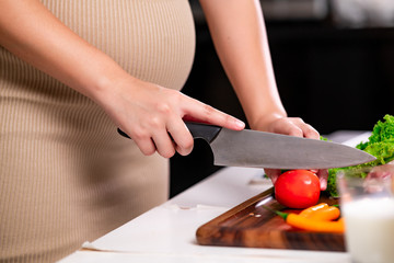Young asian pregnant woman in ketchen. Cutting tomato with large knief. Parenthood concept.